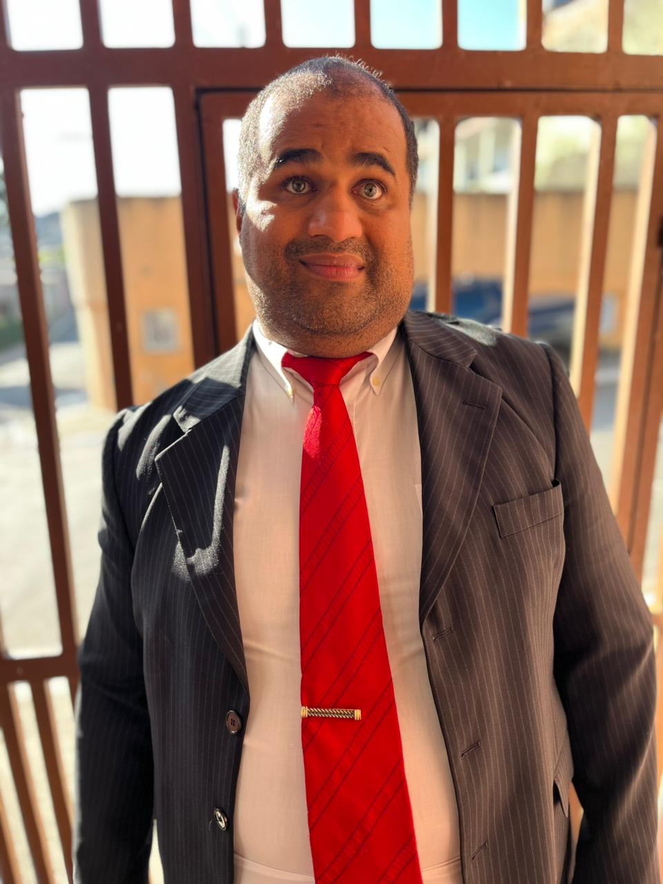 Homem de pele morena, cabelos curtos, barba por fazer e olhos esverdeados. Usa camisa branca, gravata vermelha listrada com prendedor dourado e terno preto risca de giz. Expressão neutra, com leve sorriso e iluminação lateral forte.